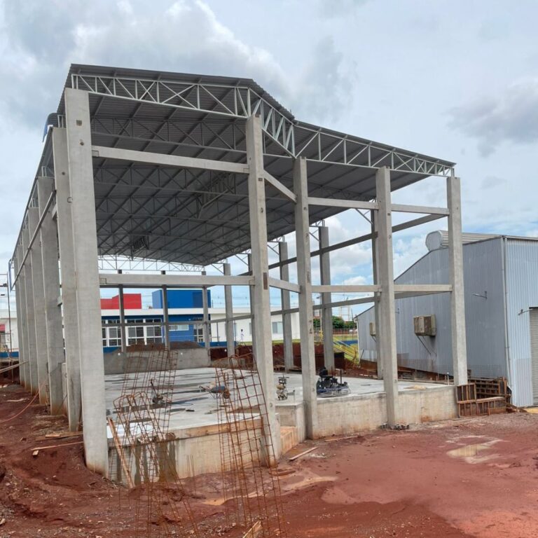Pilares e vigas de concreto pré-moldado formando a estrutura de um galpão industrial em fase inicial de construção, com fundações e área de obra ao fundo